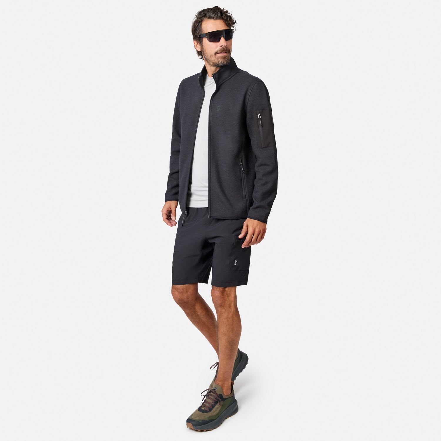 [Model is wearing size M.] A man modeling a dark gray outdoor zip-up jacket, matching dark gray shorts, a white t-shirt, and hiking shoes against a light gray background.