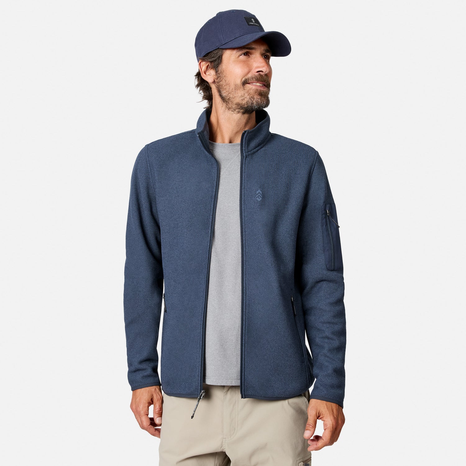 [Model is wearing size M.] A man wearing a heathered navy blue zip-up fleece jacket over a gray t-shirt, khaki pants, and a navy baseball cap, photographed against a clean light gray studio background.