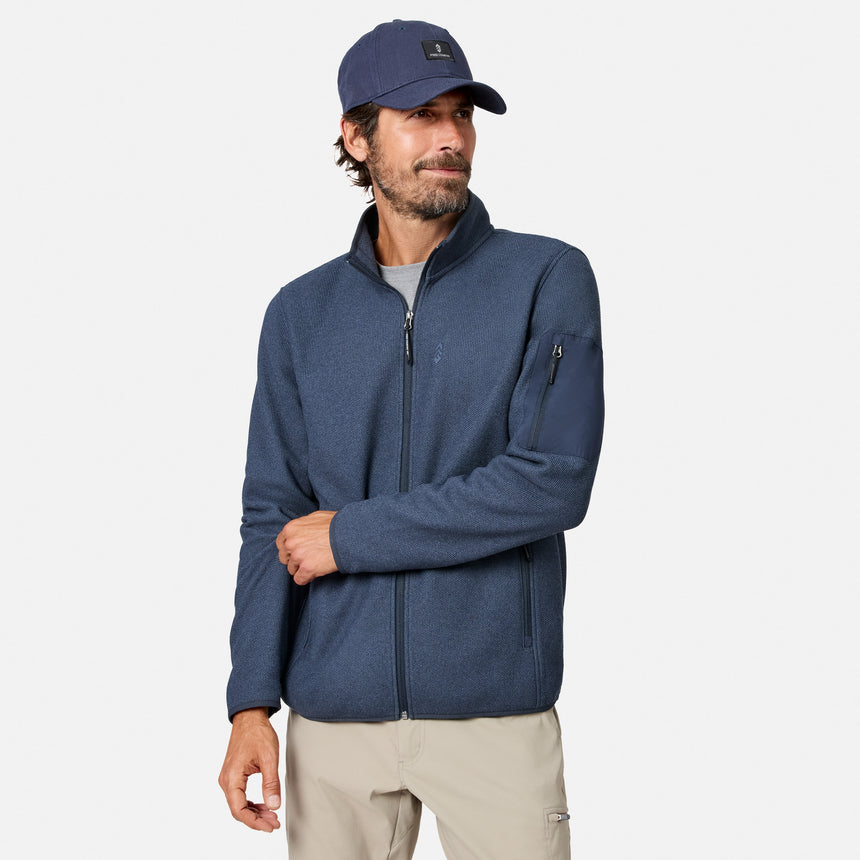 [Model is wearing size M.] A man wearing a heathered navy blue zip-up fleece jacket, a matching navy baseball cap, and beige pants, standing against a light gray studio background.