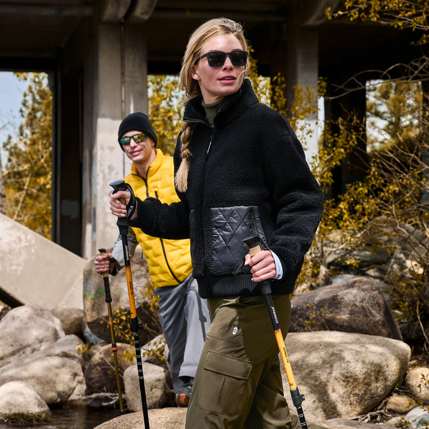 A woman in a black fleece jacket and olive cargo pants hiking with trekking poles under a concrete bridge, with a man in a yellow puffer jacket and gray pants behind her.