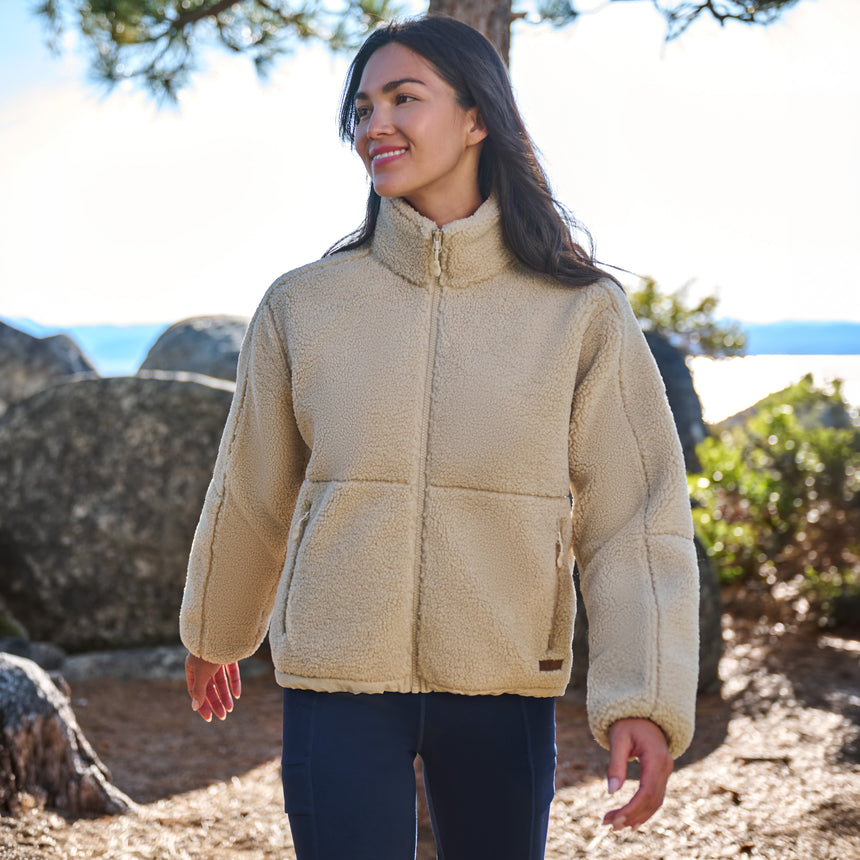 A woman wearing a beige sherpa fleece zip-up jacket with a mock neck and front pockets, standing outdoors in a rocky, wooded environment.