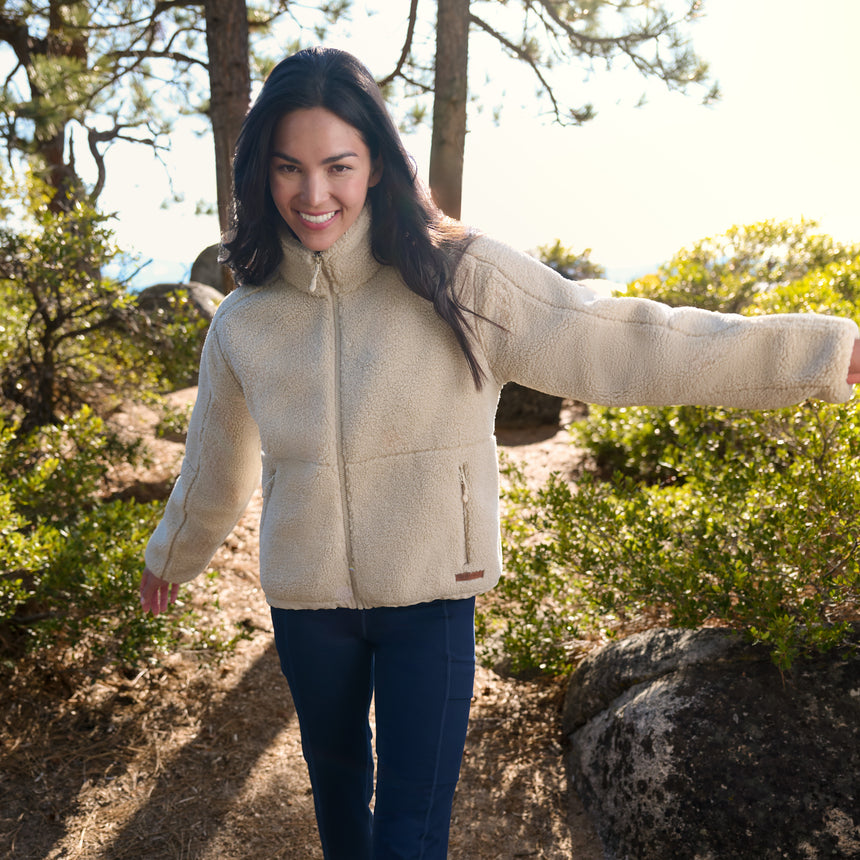 A woman wearing a cream sherpa fleece zip-up jacket and dark blue pants stands on a sunlit outdoor trail surrounded by trees and shrubs.