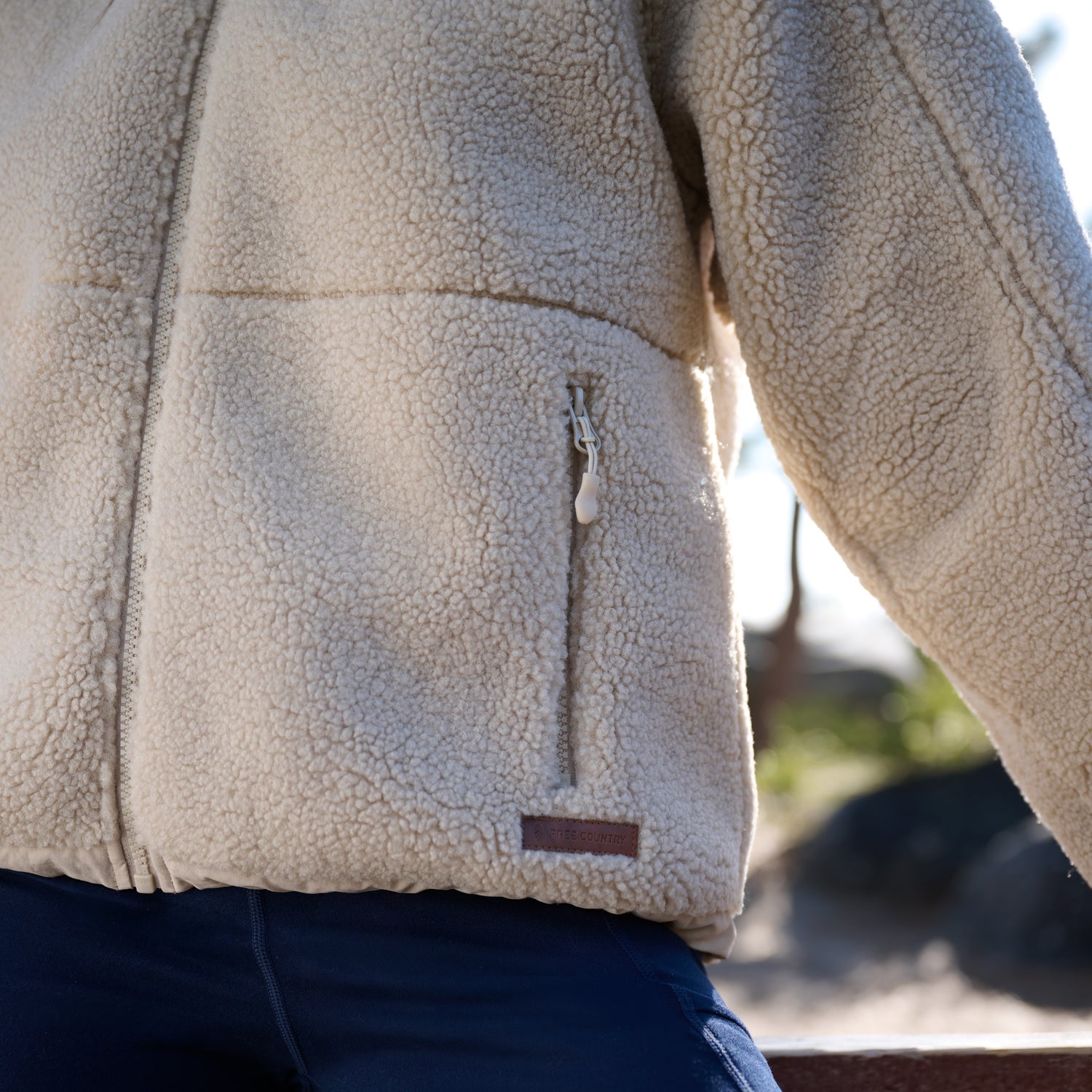 Close-up of a person wearing a cream sherpa fleece jacket with a vertical zippered pocket and a small brown brand tab, paired with dark blue pants, outdoors.