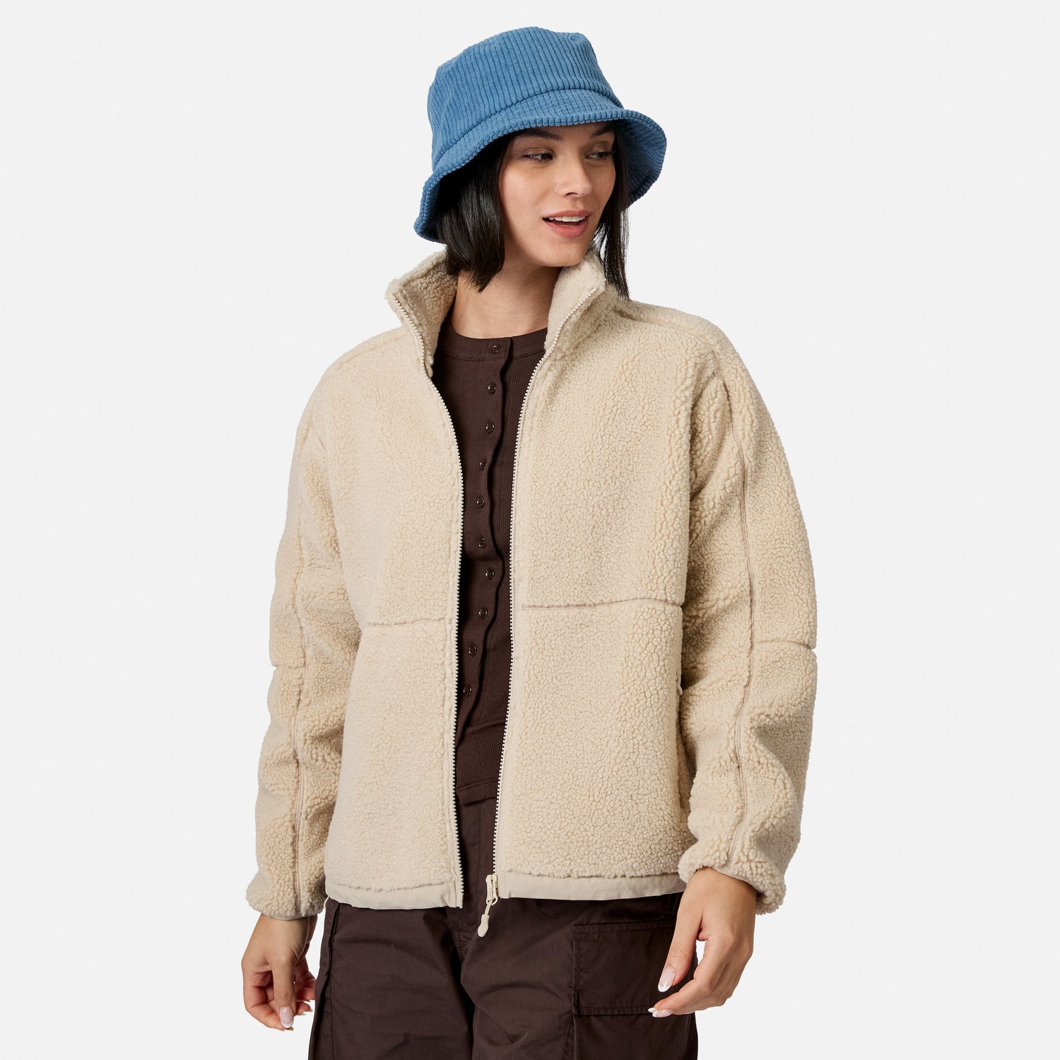 [Model is wearing size S.] A woman wearing a beige sherpa fleece zip-up jacket over a dark brown top and brown pants, paired with a blue corduroy bucket hat, against a light gray studio background.