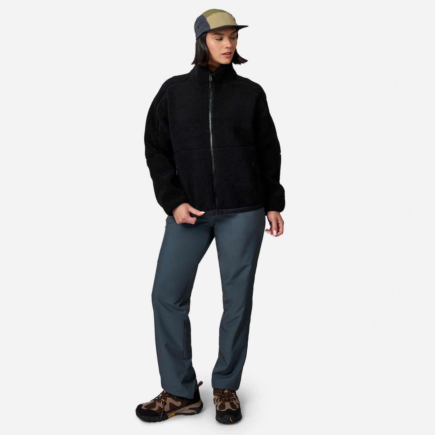 [Model is wearing size S.] A model wearing a black fleece zip-up jacket, slate blue-gray pants, brown hiking shoes, and a green and gray baseball cap, standing against a light gray background.