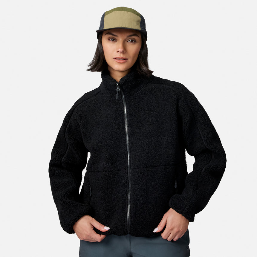 [Model is wearing size S.] A woman wearing a black fleece zip-up jacket and a green and black trucker hat, standing against a light gray studio background.