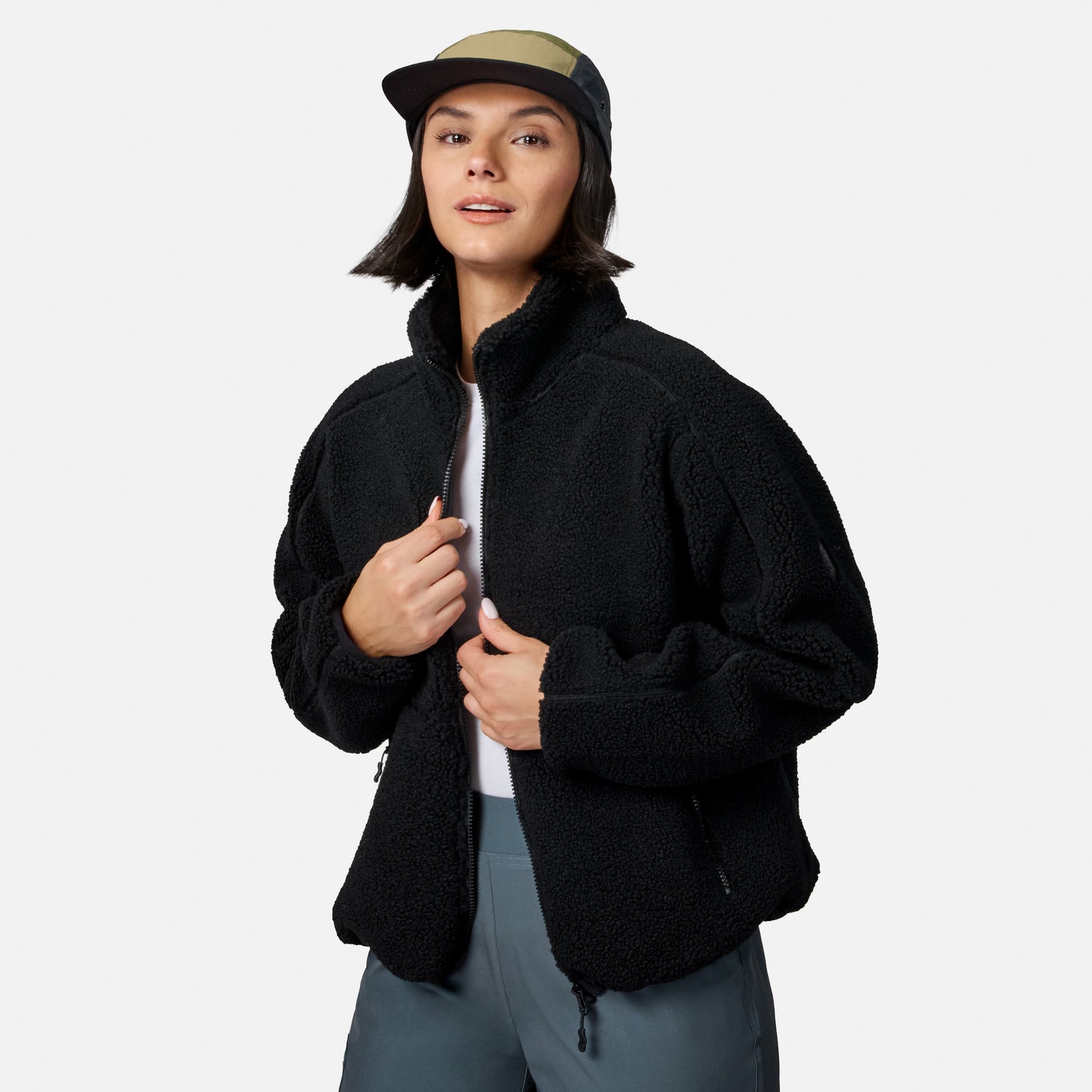 [Model is wearing size S.] A woman wearing a black sherpa fleece zip-up jacket, a black and olive green baseball cap, and gray pants, standing against a light gray studio background.