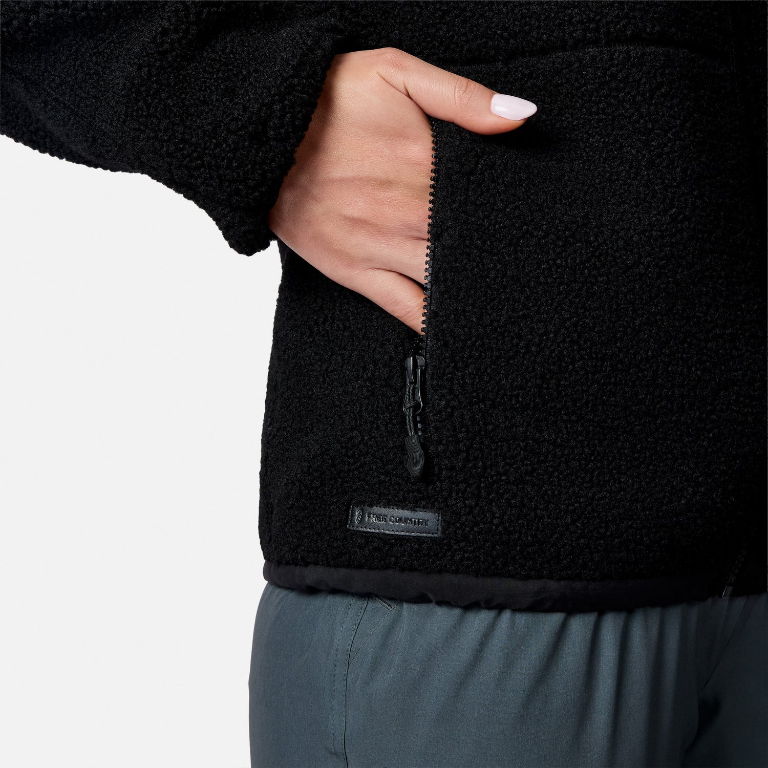 [Model is wearing size S.] Close-up of a person wearing a black fleece jacket, showing a hand in a zippered pocket and a small 'FREE COUNTRY' label near the hem, against a light gray background.