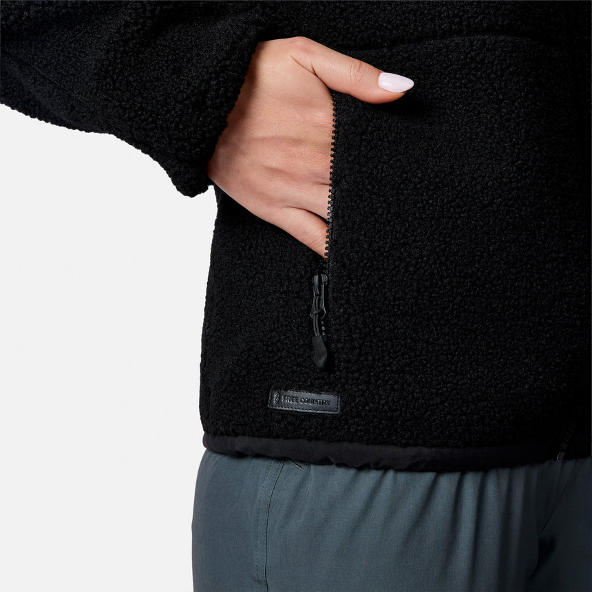 [Model is wearing size S.] Close-up of a person wearing a black fleece jacket, showing a hand in a zippered pocket and a small 'FREE COUNTRY' label near the hem, against a light gray background.