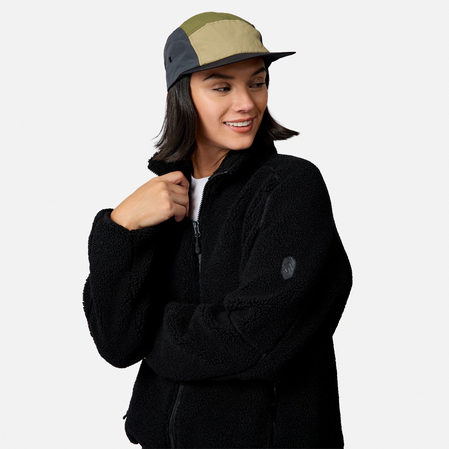 [Model is wearing size S.] A woman wearing a black fleece zip-up jacket and a multi-colored panel cap, photographed against a clean light gray studio background.