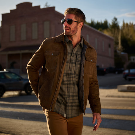 A man wearing a brown suede jacket over a green plaid flannel shirt and tan pants, walking outdoors in front of a brick building at sunset.