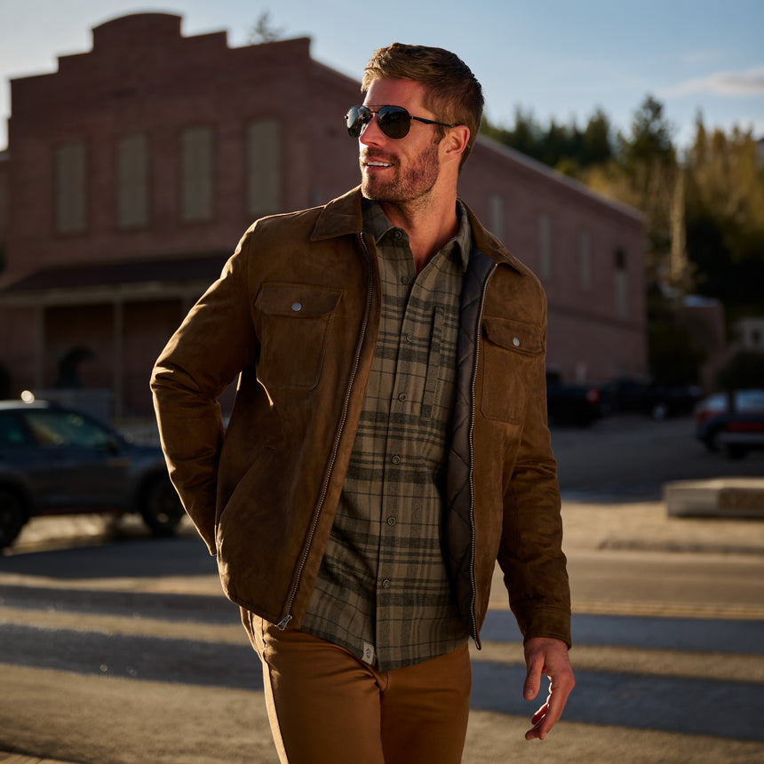 A man wearing a brown suede jacket over a green plaid flannel shirt and tan pants, walking outdoors in front of a brick building at sunset.