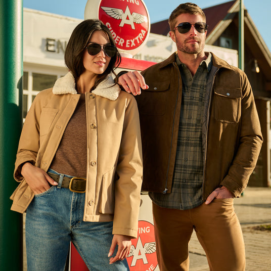 A man and a woman wearing sunglasses pose together outdoors in front of a vintage-style sign. The woman wears a tan shearling-lined jacket over a brown top and blue jeans, while the man wears a brown jacket over a green plaid shirt and brown pants.