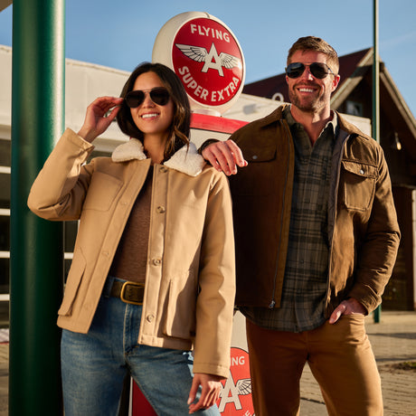 Two models wearing vintage-style aviator jackets in a retro roadside diner setting. The woman on the left wears a tan jacket with a white shearling collar, and the man on the right wears a brown jacket over a plaid shirt.