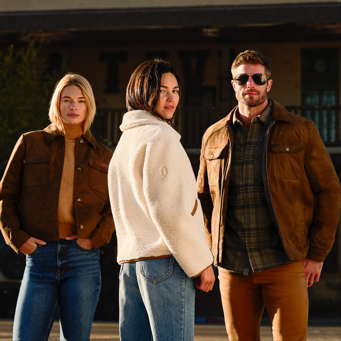 Three models wearing brown and beige outerwear stand together outdoors in front of a rustic building. The woman on the left wears a brown jacket over a tan turtleneck and blue jeans.