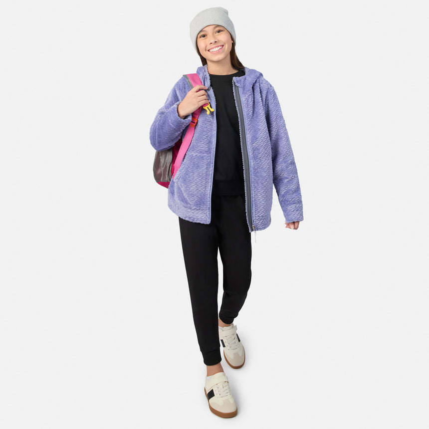 A girl wearing a grey beanie, a purple Girls’ Braided Butter Pile® Jacket, a black top, and black joggers walks forward carrying a pink backpack.