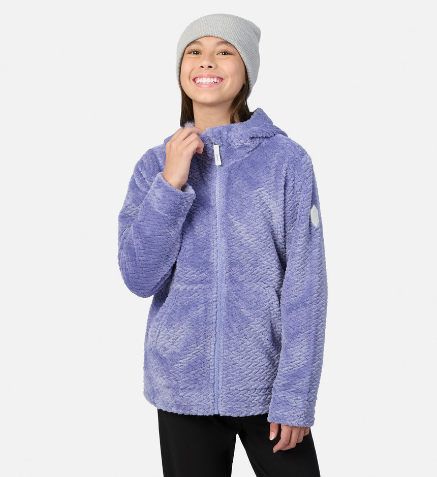 A girl smiles while wearing a grey beanie and a purple Free Country Girls’ Braided Butter Pile® Jacket, featuring a textured braided pattern and a zip-up front.
