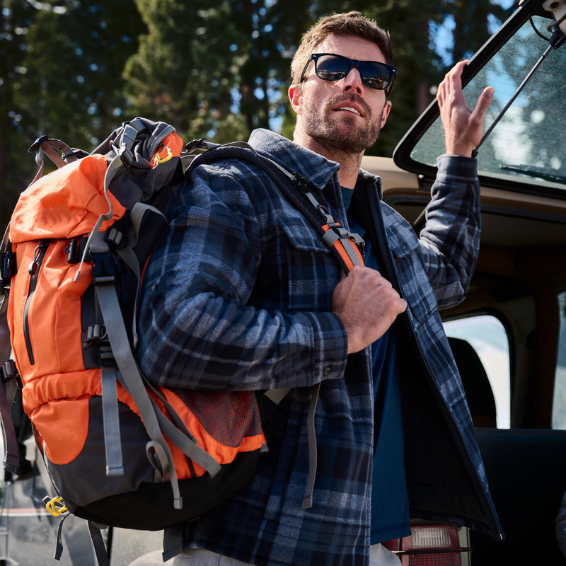 A man wearing the Free Country Men’s Chill Out Fleece Shirt Jacket and sunglasses carries an orange hiking backpack while standing next to an open car door, with trees visible in the background.