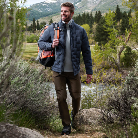 A man hiking outdoors on a trail wearing a navy blue puffer jacket over a gray shirt, brown pants, and carrying an orange and black backpack, with mountains and trees in the background.