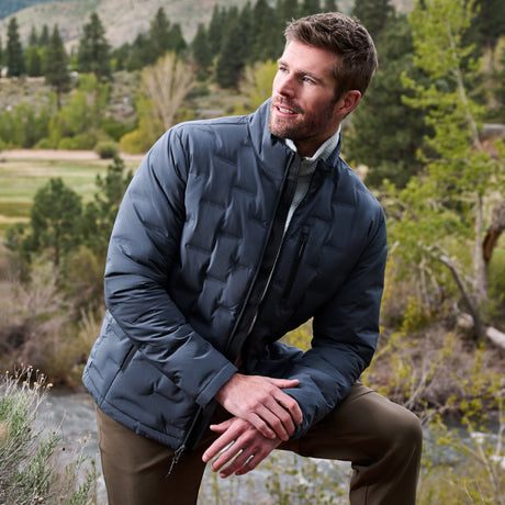 A man wearing a navy blue quilted puffer jacket and brown pants, kneeling outdoors in a natural, wooded setting.