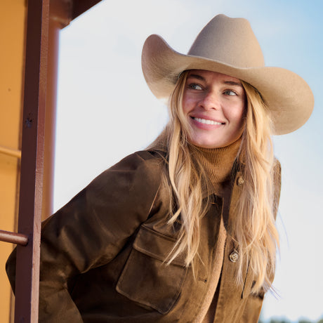 A smiling blonde woman wearing a beige cowboy hat and a brown jacket over a tan turtleneck, posed outdoors against a bright sky.