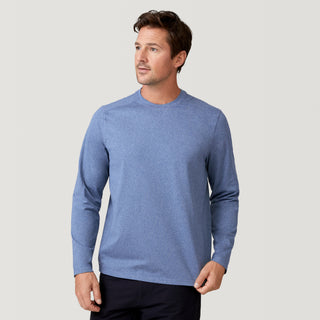 A man in the Free Country Men's FreeCycle® Sueded Melange Long Sleeve Crew Neck, light blue, pairs it with dark pants and stands against a plain light gray background, gazing slightly left with a neutral expression.