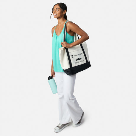 A smiling woman in a turquoise Free Country Women’s Microtech® Chill B Cool V-Neck Cami Top with Built-In Bra and white pants carries a black-and-white tote and light blue water bottle, walking against a plain white background.