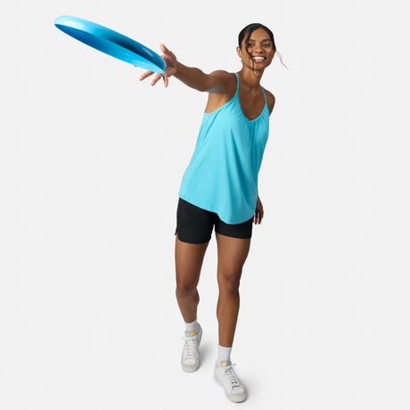 A smiling woman in a Free Country Women’s Microtech® Chill B Cool V-Neck Cami Top with Built-In Bra, black shorts, and white sneakers throws a blue frisbee on a white background, facing forward with one arm extended.