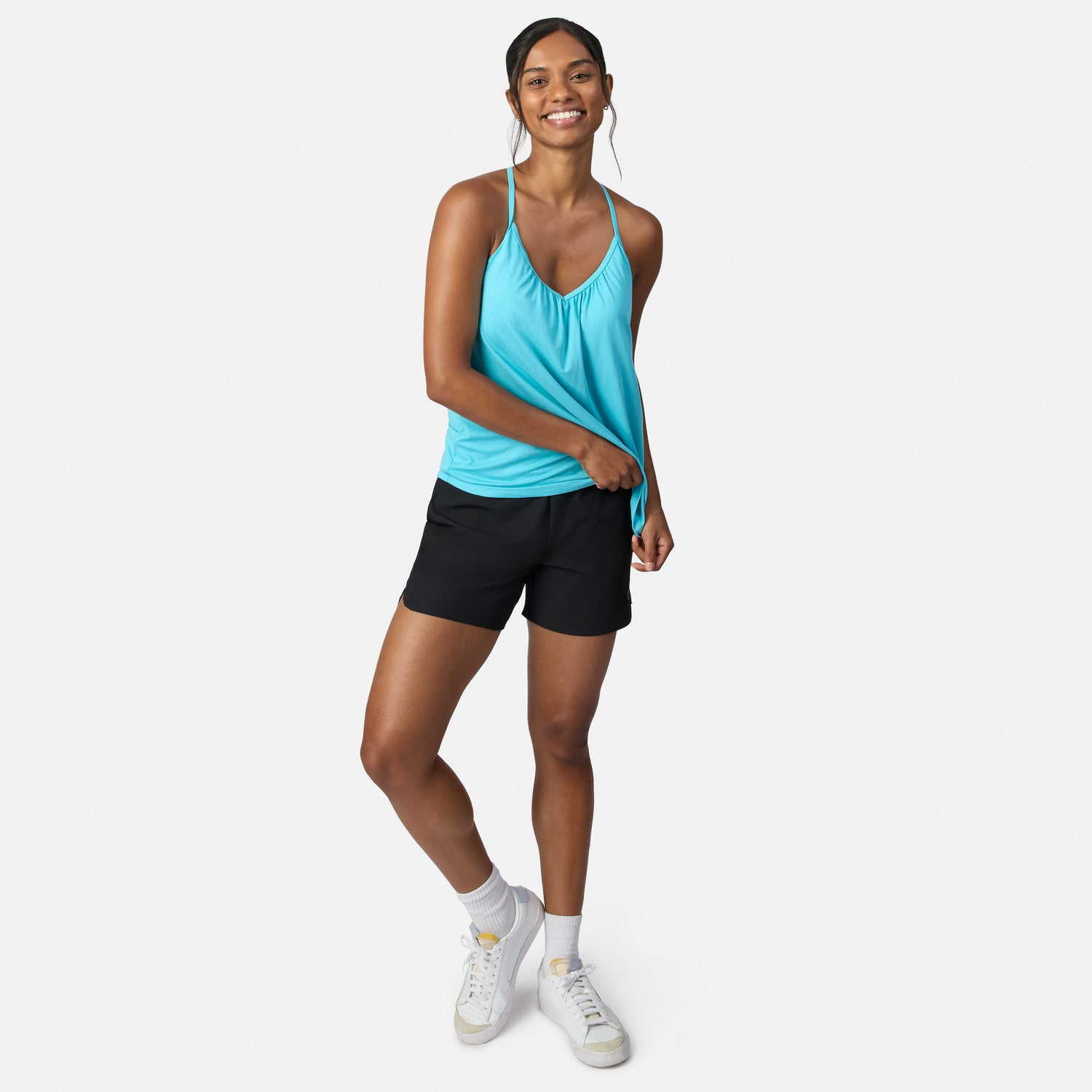 A smiling woman stands against a white background wearing a turquoise Free Country Women’s Microtech® Chill B Cool V-Neck Cami Top with Built-In Bra, black shorts, white socks, and white sneakers.