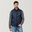 [Justin is 6’1” wearing a size Medium.] Man wearing the Free Country Men's FreeCycle® Stimson Puffer Vest in navy, featuring quilted design, REPREVE® poly fill, and a vertical zip, layered over a grey long-sleeve shirt with light jeans.