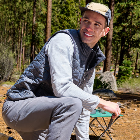 A man wearing a gray quilted vest over a light gray long-sleeve shirt and gray pants, crouching outdoors in a forest setting next to a small green folding table.