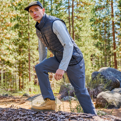 A man in a forest setting wearing a dark gray quilted puffer vest over a light gray long-sleeve shirt, dark gray pants, and tan hiking boots, crouching on a log.