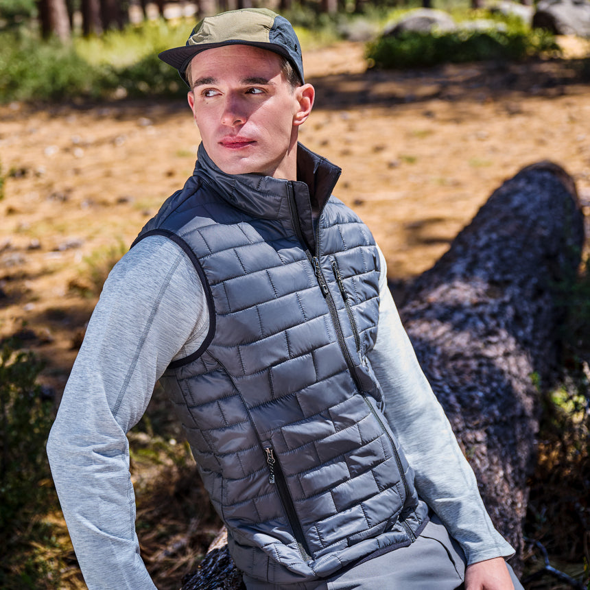 A man wearing a gray quilted puffer vest over a light gray long-sleeve shirt and a green cap, posed outdoors in a wooded setting.