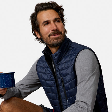 [Model is wearing size M.] A man wearing a navy blue quilted puffer vest over a gray long-sleeve shirt, holding a blue enamel mug against a plain white background.