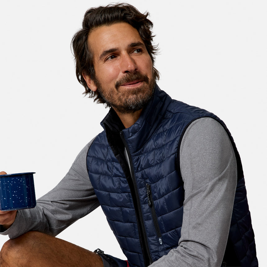 [Model is wearing size M.] A man wearing a navy blue quilted puffer vest over a gray long-sleeve shirt, holding a blue enamel mug against a plain white background.