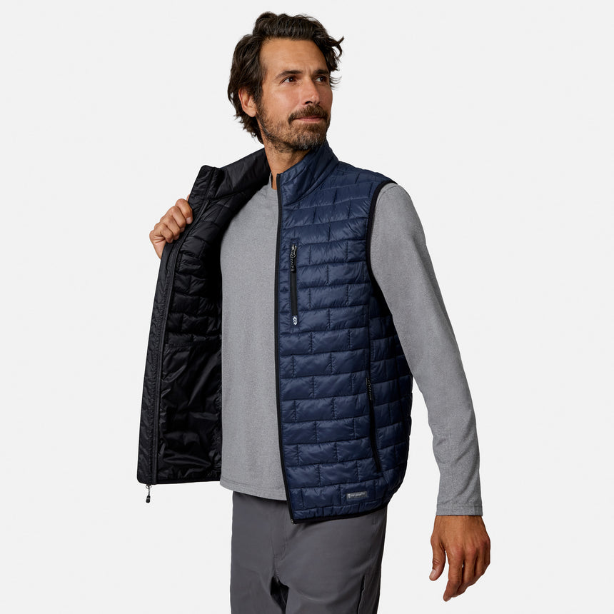 [Model is wearing size M.] A man wearing a navy blue quilted vest over a gray long-sleeve shirt and gray pants, holding the vest open to show the black interior lining, against a light gray background.