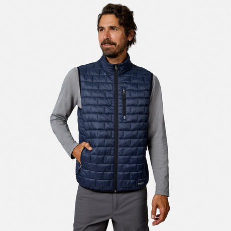 [Model is wearing size M.] A man wearing a navy blue quilted zip-up vest over a gray long-sleeve shirt and gray pants, standing against a light gray studio background.