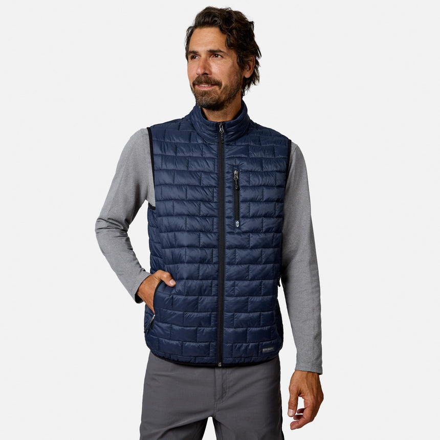 [Model is wearing size M.] A man wearing a navy blue quilted zip-up vest over a gray long-sleeve shirt and gray pants, standing against a light gray studio background.