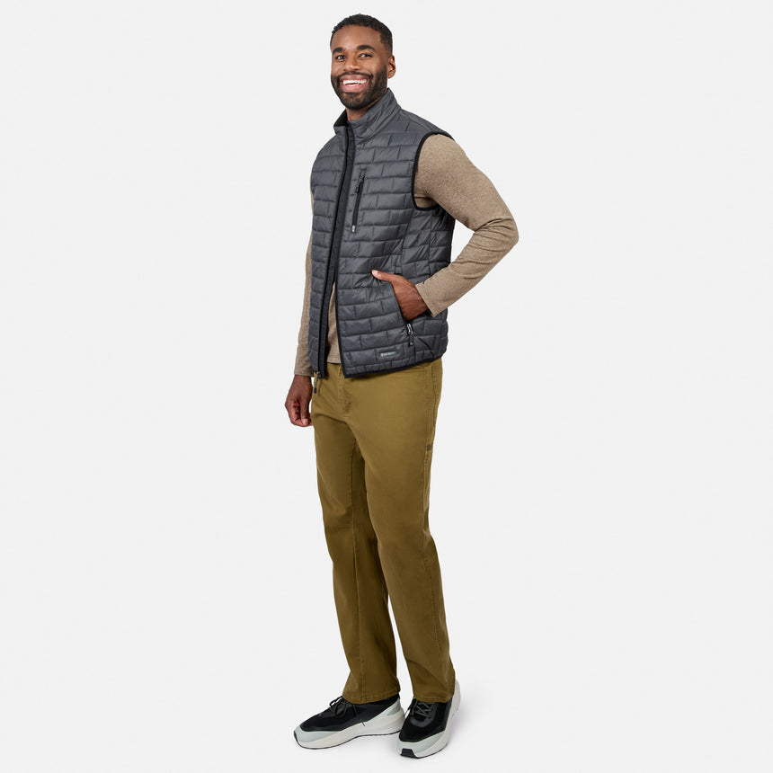 [Model is wearing size M.] A man modeling a dark gray quilted zip-up puffer vest over a beige long-sleeve shirt, olive green pants, and black sneakers, standing against a light gray studio background.