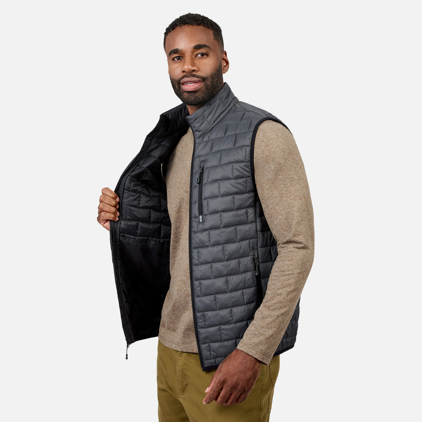 [Model is wearing size M.] A man wearing a dark gray quilted puffer vest over a beige knit sweater and olive pants, holding open the vest to show the black interior against a light gray studio background.