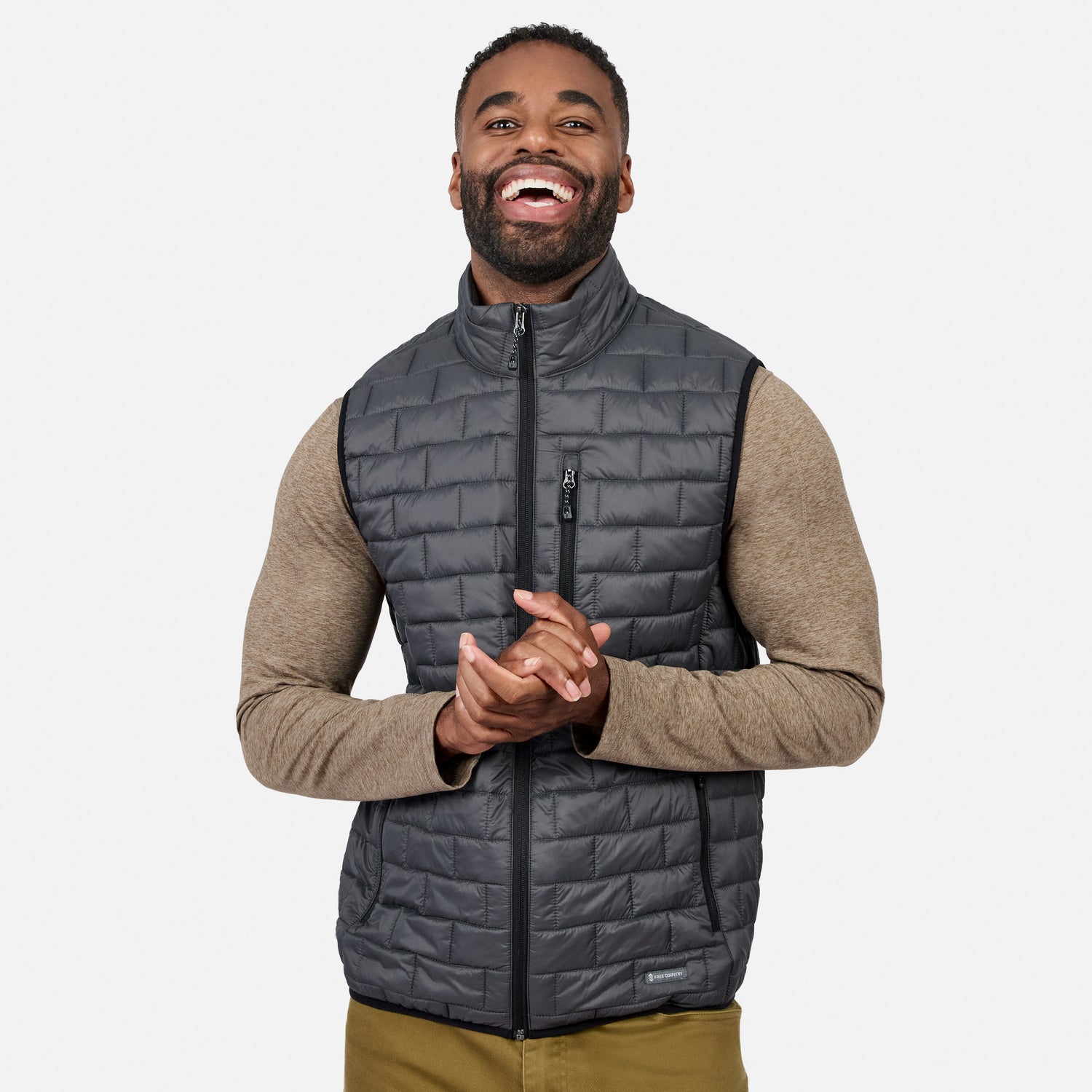 [Model is wearing size M.] A smiling man wearing a dark gray quilted zip-front puffer vest over a tan long-sleeve shirt, shown against a light gray studio background.
