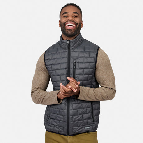 [Model is wearing size M.] A smiling man wearing a dark gray quilted zip-front puffer vest over a tan long-sleeve shirt, shown against a light gray studio background.