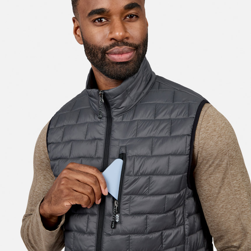 [Model is wearing size M.] A man wearing a dark gray quilted puffer vest over a beige long-sleeve shirt, holding a light blue card near the front zipper pocket.