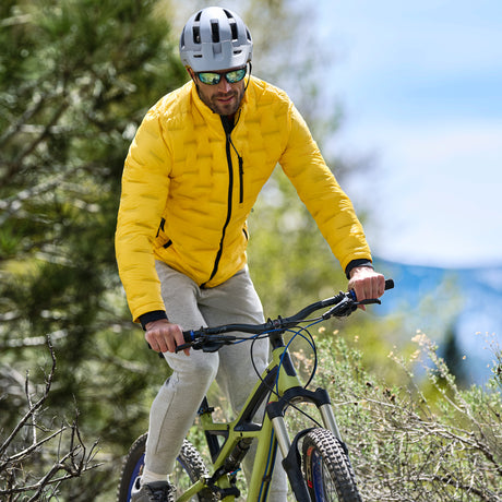 A man wearing a bright yellow puffer jacket, gray helmet, and mirrored sunglasses rides a mountain bike on a wooded trail.