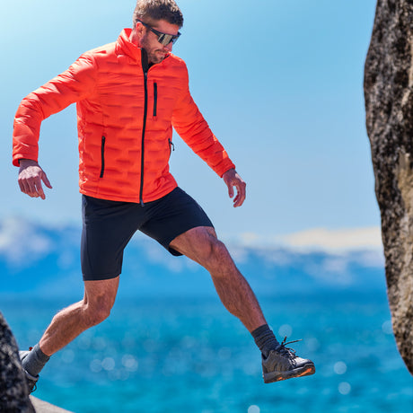 Man in a bright orange quilted jacket stepping across lakeside rocks