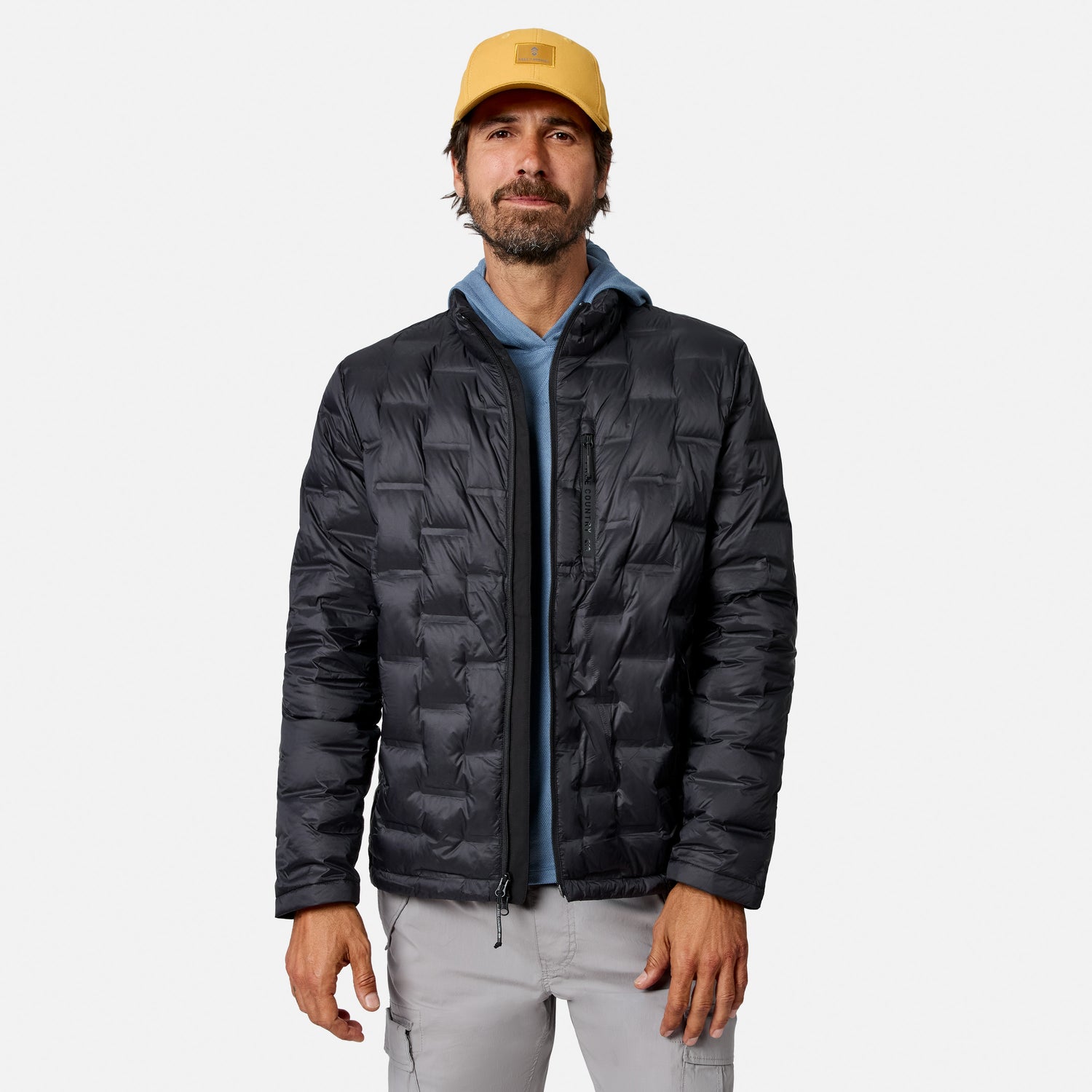 [Model is wearing size M.] A man wearing a black quilted puffer jacket over a blue hoodie, paired with a yellow cap and light gray pants, standing against a light gray studio background.
