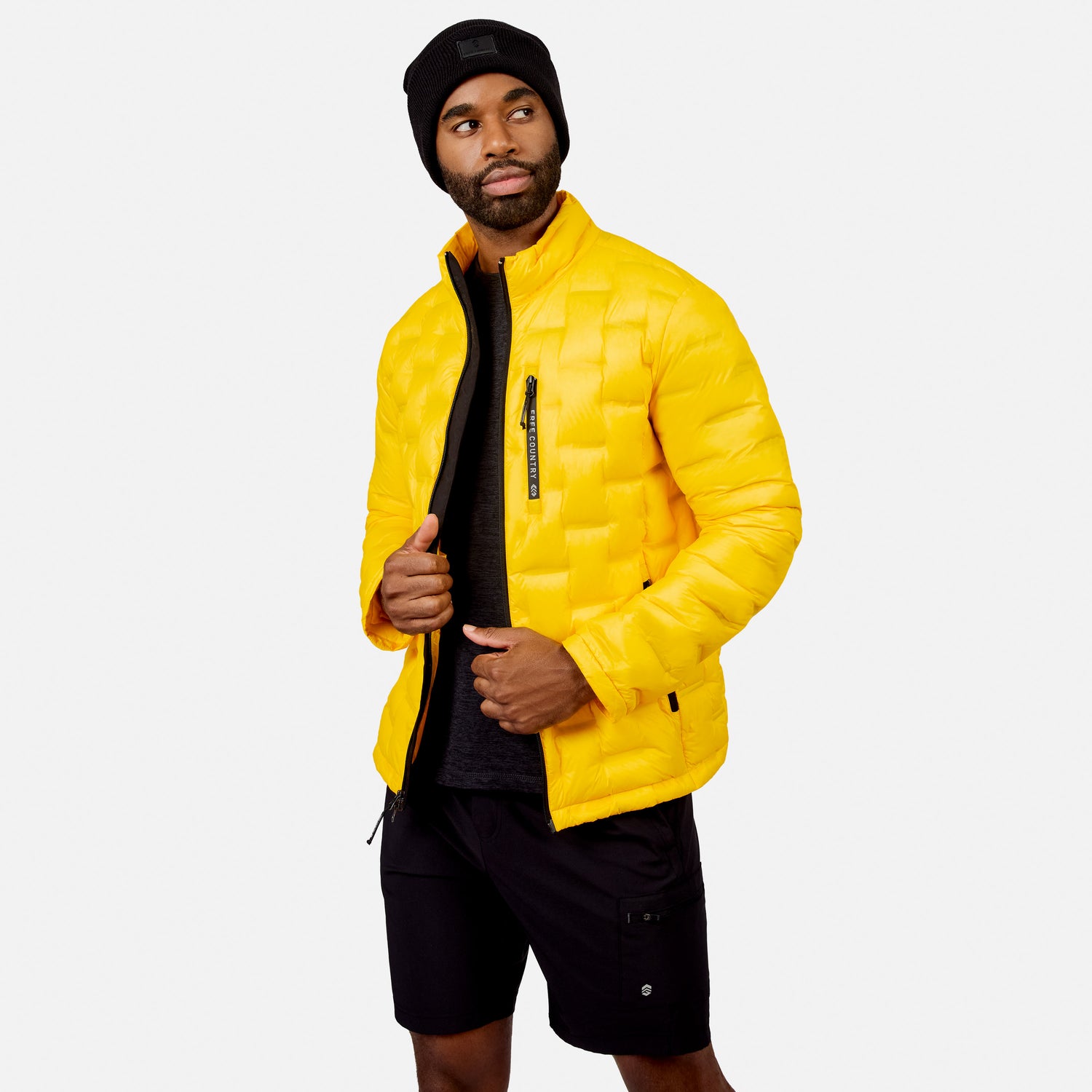 [Model is wearing size M.] A man wearing a bright yellow puffer jacket, black beanie, and black shorts, standing against a light gray studio background.