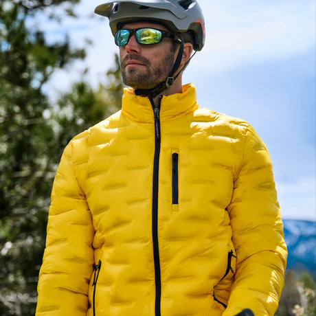 A man wearing a bright yellow puffer jacket, a gray helmet, and mirrored sunglasses, photographed outdoors against a blurred background of trees and sky.