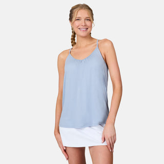A blonde woman in a braid smiles while wearing the Free Country Women's Microtech® Chill B Cool V-Neck Cami Top with Built-In Bra in light blue and a white skirt, standing against a plain white background.