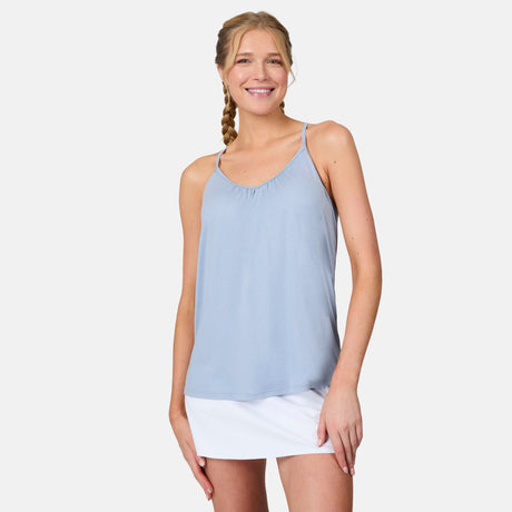 A blonde woman in a braid smiles while wearing the Free Country Women's Microtech® Chill B Cool V-Neck Cami Top with Built-In Bra in light blue and a white skirt, standing against a plain white background.